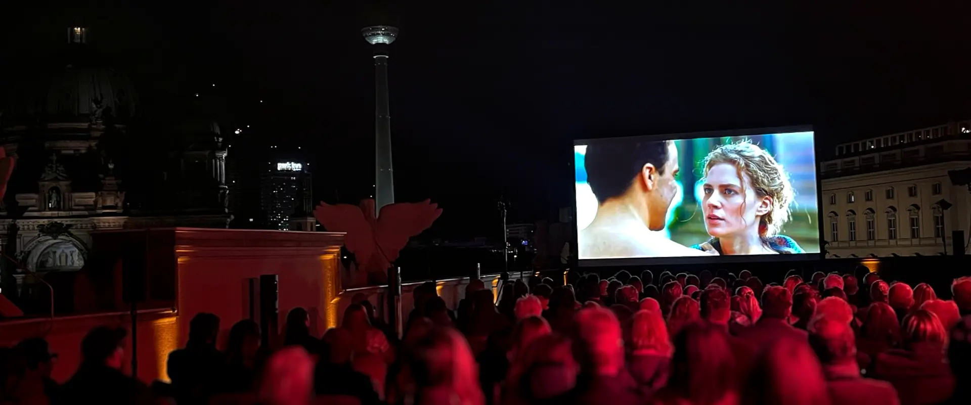 Bertelsmann Sommerkino: Blick auf die Leinwand über die Köpfe der Zuschauenden hinweg. Im Hintergrund der nächtliche Fernsehturm.
