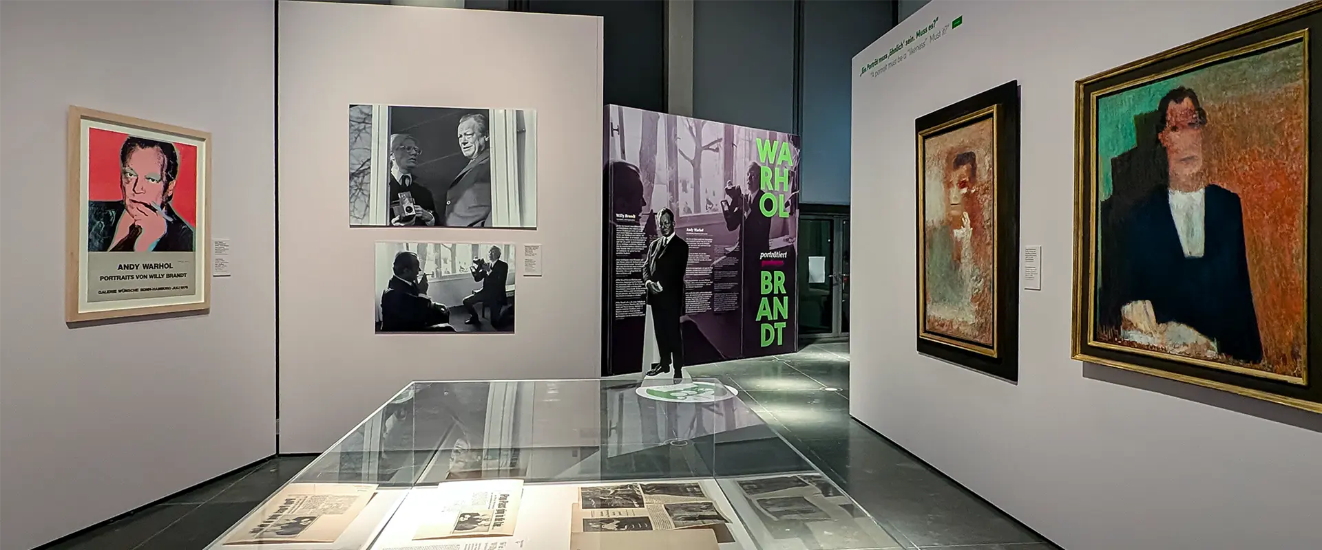 Ausstellungsraum mit mehreren Gemälden und Fotografien an den Wänden sowie Ausstellungstafeln und Vitrinen mit Dokumenten im Deutschen Bundestag zur Ausstellung „Kokoschkas Adenauer. Warhols Brandt“.