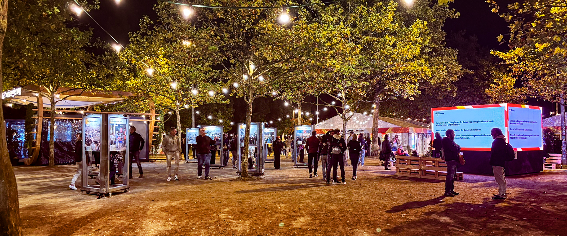 Menschen stehen und gehen unter beleuchteten Bäumen auf einem Festgelände mit Infotafeln und Lichterketten beim Tag der Deutschen Einheit 2026 in Bremen.